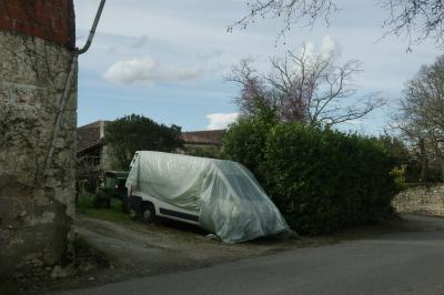 photograph “III.2025” par David Farreny — www.farreny.net — France, Occitanie, Gascogne, Gascony, Lomagne, Gers, Monfort, village, fourgonnette, van, bâche, plastique, plastic, tarpaulin, protection, véhicule, vehicle, gouttière, drainpipe, bâtiment, building, pierre, stone, parpaings, breeze-blocks, briques, bricks, métal, metal, rue, street, bitume, blacktop, asphalte, asphalt, nuages, clouds, ciel, sky, haie, hedge, arbres, trees, tracteur, tractor, phares, headlights, maisons, houses, toits, roofs, tuiles, tiles, mur, wall, branches