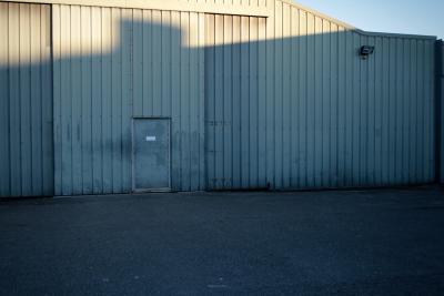 photograph “XII.2011” par David Farreny — www.farreny.net — France, entrepôt, warehouse, tôle ondulée, corrugated iron, métal, metal, industrie, industry, façade, facade, porte, door, projecteur, light, portail, gate, bitume, blacktop, asphalte, asphalt, soir, evening, ombre, shadow, vide, empty, emptiness, Occitanie, Aveyron, Rouergue, Olemps