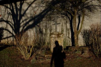 photograph “II.2015” par David Farreny — www.farreny.net — France, autoportrait, selfportrait, ombre, shadow, silhouette, croix, cross, pierre, stone, métal, metal, socle, plinth, pedestal, arbres, trees, ombres, shadows, mur, wall, parpaings, breeze-blocks, branches, soir, evening, arbustes, shrubberies, herbe, grass, profusion, Occitanie, Aveyron, Rouergue, Olemps