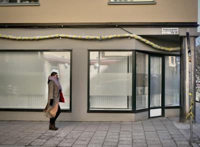 photograph “II.2022 — Suspension” par David Farreny — www.farreny.net — Suède, Sverige, Sweden, Uppland, Södermanland, Stockholm, ville, city, capitale, capital, hiver, winter, immeuble, bâtiment, building, femme, woman, passante, passer-by, piétonne, pedestrian, vitrine, window, bay, verre, vitre, glass, trottoir, pavement, rue, street, Högbergsgatan, 75A-85, plaque, plate, pilier, pillar, coin, angle, corner, reflet, reflection, panneau, sign, routier, roadsign, voiture, car, écharpe, scarf, lunettes, glasses, manteau, coat, marche, walk, stores, tuyau, pipe, hachures, hatching, jaune, yellow, ruban, ribbon, fenêtres, windows