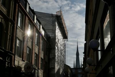 photograph “II.2022 — Run away to the North, 216” par David Farreny — www.farreny.net — Suède, Sverige, Sweden, Uppland, Södermanland, Stockholm, ville, city, capitale, capital, immeubles, bâtiments, buildings, rue, street, fenêtres, windows, hiver, winter, soleil, sun, reflet, reflection, travaux, works, échafaudage, scaffolding, église, church, clocher, tower, nuages, clouds, Frantzén, lampes, lamps, métal, metal, croix, cross, Norrmalm
