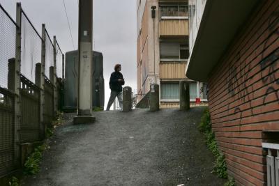 photograph “III.2024” par David Farreny — www.farreny.net — France, Occitanie, Haute-Garonne, Toulouse, ville, city, rue, street, ruelle, alley, passage, asphalte, asphalt, bitume, blacktop, mur, wall, brique, brick, béton, concrete, immeuble, bâtiment, building, clôture, fence, poteaux, posts, poles, balcon, balcony, fenêtres, windows, passant, passer-by, piéton, pedestrian, homme, man, jeune, young, conteneur, container, plastique, plastic, panneau, sign, tags, graffitis, câbles, cables, fils, wires