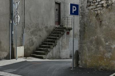 photograph “VII.2020 — Faces of home, 111” par David Farreny — www.farreny.net — France, Auvergne, Rhône-Alpes, Haute-Loire, Le Monastier-sur-Gazeille, ville, city, rue, street, carrefour, crossing, bitume, blacktop, asphalte, asphalt, panneau, routier, sign, roadsign, P, lettre, letter, parking, car park, gouttière, drainpipe, câbles, cables, fils, wires, maison, house, escalier, stairs, marches, steps, pierre, stone, ciment, cement, porte, door, bois, wood, métal, metal, coin, corner, angle, plaque, plate, domicile, home, gris, grey, gray