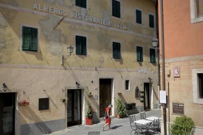 photograph “VIII.2022” par David Farreny — www.farreny.net — Italie, Italia, Italy, Toscane, Toscana, Tuscany, Sienne, Siena, San Quirico d'Orcia, rue, street, bâtiment, immeuble, building, angle, coin, corner, fenêtres, windows, portes, doors, tables, chaises, chairs, tuyau, pipe, femme, woman, passante, passer-by, piétonne, robe, dress, rouge, red, lampes, lamps, briques, bricks, volets, shutters, bois, wood, panneaux, signs, enseigne, Albergo ristorante L'Orcia, façade, facade, pierre, stone, ville, city, bourgade, town