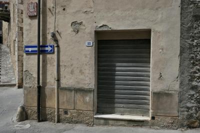 photograph “VIII.2022” par David Farreny — www.farreny.net — Italie, Italia, Italy, Toscane, Toscana, Tuscany, Sienne, Siena, San Quirico d'Orcia, rue, street, coin, corner, angle, bâtiment, building, pierre, stone, gouttière, drainpipe, métal, metal, panneau, sign, flèche, arrow, câble, cable, fil, wire, mur, wall, balcon, balcony, porte, door, numéro, number, 35, Castello