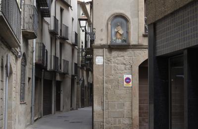 photograph “VI.2023” par David Farreny — www.farreny.net — Espagne, España, Espanya, Spain, Catalogne, Cataluña, Catalunya, Catalonia, Lérida, Lleida, Solsonès, Solsona, ville, city, centre ancien, old center, carrefour, intersection, crossroads, maisons, houses, bâtiments, buildings, pierre, stone, angle, coin, corner, ruelle, alley, rue, street, fenêtres, windows, balcons, balconies, lampe, lamp, lampadaire, métal, metal, panneau, sign, portes, doors, gouttière, drainpipe, plaque, plate, travessia dels Dominics, statue, sculpture, christianisme, Christianism, catholicisme, Catholicism, croix, cross, fleurs, flowers