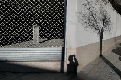 photograph “I.2023” par David Farreny — www.farreny.net — Espagne, España, Espanya, Spain, Catalogne, Cataluña, Catalunya, Lleida, Lérida, Alt Urgell, La Seu d'Urgell, ville, city, rue, street, angle, coin, corner, bâtiment, immeuble, building, béton, concrete, trottoir, pavement, pierre, stone, grille, fencing, métal, metal, rideau, curtain, bloc, block, arbre, tree, ombre, shadow, autoportrait, selfportrait, photographe, photographer