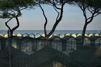 photograph “V.2022” par David Farreny — www.farreny.net — Italie, Italia, Italy, Vénétie, Veneto, Venise, Venice, Venezia, Venexia, Lido, mer, sea, plage, beach, arbres, trees, pins, pines, cabines, cabins, bois, wood, grillage, fence, clôture, fencing, métal, metal, toits, roofs, eau, water, bateaux, boats, navires, ships, horizon, ciel, sky, troncs, trunks, drapeau, flag