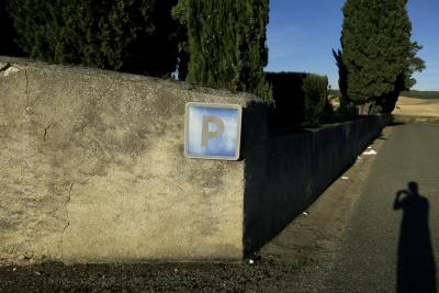 photograph “XI.2015” par David Farreny — www.farreny.net — France, Occitanie, Gascogne, Gascony, Gers, Jegun, cimetière, cemetery, mur, wall, pierre, stone, enduit, plaster, coin, corner, angle, panneau, sign, P, parking, lettre, letter, bleu, blue, cyprès, cypresses, arbres, trees, route, road, bitume, blacktop, asphalte, asphalt, ombre, shadow, autoportrait, selfportrait, collines, hills