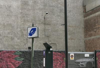 photograph “IX.2023 — Whatever” par David Farreny — www.farreny.net — Andorre, Andorra, Andorre-la-Vieille, Andorra la Vella, ville, city, immeuble, building, bâtiment, mur, wall, brique, brick, béton, concrete, ciment, cement, crépi, roughcast, peinture, painting, parking, car park, poteau, post, pole, métal, metal, lampes, lampadaire, lamps, rue, street, panneau, sign, horodateur, parking meter, logo, main, hand, pièce, coin, câble, cable, fil, wire, angle, corner, clôture, fence, barrière, voiture, car, fleurs, flowers, rouge, red