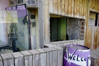 photograph “VII.2023 — Welcome” par David Farreny — www.farreny.net — Andorre, Andorra, Encamp, Le Pas de la Case, El Pas de la Casa, immeuble, bâtiment, building, restaurant, terrasse, terrace, mur, paroi, wall, bois, wood, pierre, stone, verre, vitre, glass, table, chaises, chairs, planches, planks, fenêtre, window, grille, railings, tonneau, barrique, barrel, welcome, bienvenue, plats du jour, daily specials, inscriptions, signs, violet, purple, peinture, paint