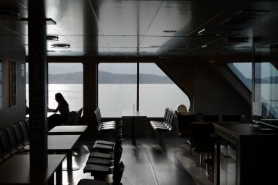 photograph “V.2019 — Run away to the North, 172” par David Farreny — www.farreny.net — Norvège, Norway, Norge, Vestlandet, Hordaland, Bergen, Krokeide, ferry, bateau, boat, bac, Hufthamar, Austevoll, Lysefjord, fjord, mer, sea, eau, water, collines, hills, baie, fenêtre, window, verre, glass, vitre, chaises, chairs, tables, métal, metal, pont, deck, soir, evening, été, summer, vide, empty, emptiness, femme, woman, jeune femme, young woman, plafond, ceiling, paysage, landscape, marin, seascape