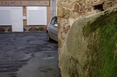 photograph “II.2014” par David Farreny — www.farreny.net — France, Occitanie, Aveyron, Rouergue, Cassagnes-Bégonhès, village, place, square, rue, street, bitume, asphalte, asphalt, blacktop, voiture, car, rétroviseur, wing mirror, magasin, store, boutique, shop, enseigne, sign, fermé, closed down, Calmels, mercerie, chapellerie, haberdashery, hat shop, porte, door, fenêtres, windows, volets, stores, gouttière, drainpipe, métal, metal, mousse, moss, pierre, stone, église, church, mosaïque, mosaic, jardinières, planters, pots