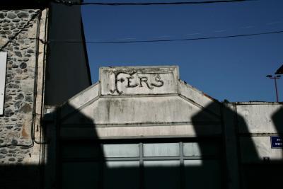 photograph “III.2012 — Irons” par David Farreny — www.farreny.net — France, Occitanie, Aveyron, Rouergue, Espalion, bâtiment, building, maison, house, atelier, workshop, enseigne, sign, fers, irons, pierre, stone, béton, concrete, câbles, cables, fils, wires, volet, shutter, bois, wood, ombre, shadow, lampadaire, lamp, plaque, plate, avenue d'Estaing, numéro, number, 2, ciel, sky