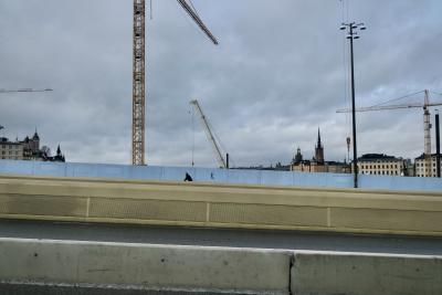 photograph “II.2022 — Run away to the North, 203” par David Farreny — www.farreny.net — Suède, Sverige, Sweden, Uppland, Södermanland, Stockholm, ville, city, capitale, capital, pont, bridge, route, road, béton, concrete, muret, low wall, bâtiments, buildings, grues, cranes, chantier, works, construction, nuages, clouds, hiver, winter, câbles, cables, métal, metal, homme, man, cycliste, cyclist, clocher, bell tower, tour, tower, bitume, blacktop, asphalte, asphalt, palissade, palisade, fence, bleu, blue, lampadaire, lamp, fenêtres, windows