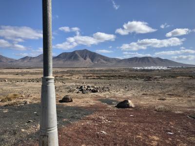 photograph “II.2018 — Lancerotte, 78” par David Farreny — www.farreny.net — Espagne, Spain, España, Canaries, Canarias, Lanzarote, Lancerotte, Yaiza, Playa Blanca, île, island, plaine, plain, montagnes, mountains, poteau, post, pole, métal, metal, lampadaire, lamp, rochers, rocks, ciel, sky, nuages, clouds, maisons, houses, blanches, white, paysage, landscape, sol, soil, terre, earth, rouge, red, jaune, yellow