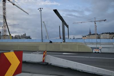 photograph “II.2022 — Run away to the North, 280” par David Farreny — www.farreny.net — Suède, Sverige, Sweden, Uppland, Södermanland, Stockholm, ville, city, capitale, capital, pont, bridge, route, road, béton, concrete, murets, low walls, bâtiments, buildings, grues, cranes, chantier, works, construction, nuages, clouds, hiver, winter, métal, metal, homme, man, cycliste, cyclist, clocher, bell tower, bitume, blacktop, asphalte, asphalt, palissade, palisade, fence, bleu, blue, lampadaire, lamp, fenêtres, windows, panneaux, signs, routiers, roadsigns, flèches, arrows, buss
