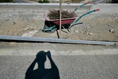 photograph “VII.2023 — Self-portrait with electric fagot” par David Farreny — www.farreny.net — Andorre, Andorra, Canillo, route, road, bitume, blacktop, asphalte, asphalt, chantier, works, trottoir, pavement, béton, concrete, métal, metal, poteau, post, pole, bois, wood, brouette, wheelbarrow, armatures, treillis, trellis, roue, wheel, poignées, handles, ombre, shadow, photographe, photographer, terre, earth, sol, soil, cailloux, pierres, stones, pebbles