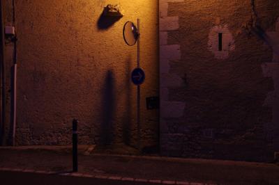 photograph “V.2016” par David Farreny — www.farreny.net — France, Occitanie, Gascogne, Gers, Jegun, rue, street, nuit, night, soir, evening, mur, wall, pierre, stone, crépi, roughcast, poteau, pole, post, métal, metal, miroir, mirror, trottoir, pavement, ouverture, opening, lumière, light, panneau, sign, roadsign, flèche, arrow, droite, right, câbles, cables, fils, wires, boîtier, box, goulotte, chute, coin, angle, corner