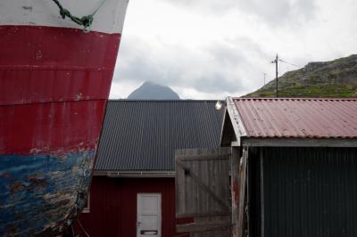 photograph “VII.2017 — Run away to the North, 119” par David Farreny — www.farreny.net — Norvège, Norway, Norge, Norvège du Nord, Nord-Norge, Nordland, Flakstad, Sund, Lofoten, bateau, boat, ship, pêche, fishing, port, harbour, village, bâtiments, buildings, métal, metal, corde, rope, proue, bow, prow, tôle ondulée, corrugated iron, toits, roofs, porte, door, bois, wood, montagne, mountain, nuages, clouds, poteaux, électriques, electricity, poles, rocher, rock, lampe, lamp, lumière, light