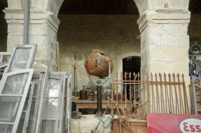 photograph “II.2025 — Spotlight on mess” par David Farreny — www.farreny.net — France, Occitanie, Gascogne, Gascony, Lomagne, Gers, Lectoure, antiquités, brocante, antiques, flea market, Village des Brocanteurs, Second-hand dealers' Village, vieux, objets, old, objects, portail, gate, métal, metal, rouille, rust, rusty, piliers, pillars, arcade, arche, arch, pierre, stone, beige, bois, wood, panneau, publicité, pub, panel, advertising, ad, Esso, logo, portes, doors, fenêtres, windows, projecteur, floodlight, poignées, handles, tuyau, pipe, gouttière, drainpipe, verre, vitre, glass, reflet, reflection, arbre, tree, branches, branchages, table, fauteuil, sofa, strates, time, temps, passé, past, profusion