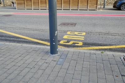 photograph “VI.2024” par David Farreny — www.farreny.net — Andorre, Andorra, Canillo, rue, street, route, road, trottoir, pavement, pierre, stone, dallage, paving, bitume, blacktop, asphalte, asphalt, poteau, post, pole, métal, metal, peinture, paint, inscription, bus, arrêt, stop, jaune, yellow, red, rouge, voiture, car, chaises, chairs, palissade, bois, wood, paroi, wall