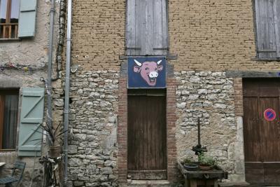 photograph “I.2024 — Joy that lasts” par David Farreny — www.farreny.net — France, Occitanie, Tarn-et-Garonne, Montricoux, bourgade, town, village, rue, street, maison, house, façade, facade, vieille, old, brique, crue, raw, brick, beige, pierre, stone, porte, door, fenêtres, windows, volets, shutters, bois, wood, portail, gate, pots, plants, plantes, gouttières, drainpipes, métal, metal, câbles, cables, fils, wires, guirlandes, Noël, Christmas, wreaths, boules, balls, chaise, chair, enseigne, sign, La Vache qui rit, The Laughing Cow, fromage, cheese, vache, cow, rouge, red, rire, laughter, Benjamin Rabier, publicité, advertising, pub, ad, panneau, ne pas stationner, do not park