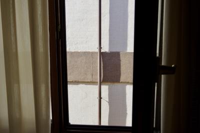 photograph “VII.2019” par David Farreny — www.farreny.net — Espagne, Spain, España, Asturies, Asturias, Llanes, hôtel, hotel, chambre, room, mur, wall, cadre, frame, fenêtre, window, rideau, curtain, beige, poignée, handle, métal, metal, carrelage, tiling, tuyau, pipe, vertical, ombre, shadow
