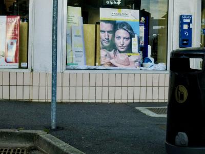 photograph “VII.2012 — The pursuit of happiness” par David Farreny — www.farreny.net — France, Occitanie, Aveyron, Rouergue, Rodez, rue, street, trottoir, pavement, bitume, blacktop, asphalte, asphalt, pharmacie, pharmacy, drugstore, vitrine, window, baie, bay, vitre, verre, glass, poubelle, bin, trash, can, poteau, post, pole, métal, metal, carrelage, tiling, panneaux, boards, publicité, advertising, pub, ad, crème solaire, solar cream, La Roche-Posay, couple, homme, man, femme, woman, jeune, young, blonde, belle, beautiful, mannequin, modèle, model, chapeau, hat, paille, straw, distributeur, préservatifs, condom, dispenser, machine, capotes, Manix, reflet, reflection
