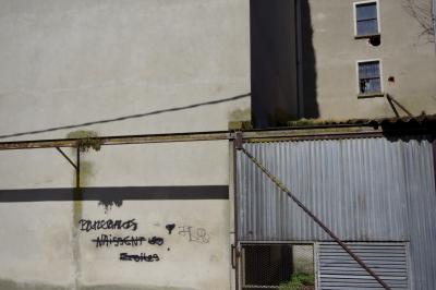 photograph “III.2021” par David Farreny — www.farreny.net — France, Occitanie, Gascogne, Gers, Fleurance, usine, factory, mur, wall, ciment, cement, tôle ondulée, corrugated iron, métal, metal, portail, gate, toit, roof, fenêtres, windows, grillage, fence, fencing, tag, graffiti, Du chaos naissent les étoiles, Stars are born from chaos, barré, strikethrough, rouille, rust, rusty, angle, coin, gouttière, drainpipe, ombre, shadow, gris, grey, gray