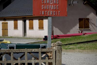 photograph “VII.2023 — Subliminal message” par David Farreny — www.farreny.net — France, Occitanie, Ariège, Mérens-les-Vals, maison, house, clôture, fence, ciment, cement, panneau, sign, métal, metal, camping sauvage interdit, wild camping prohibited, rouge, red, tuyaux, pipes, volets, shutters, bois, wood, porte, door, antenne, antenna, conteneurs, poubelle, trash, containers, plastique, plastic, herbe, grass, toit, roof, ardoise, slate