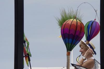 photograph “VI.2015 — Passing summer” par David Farreny — www.farreny.net — France, Aquitaine, Guyenne, Gascogne, Gascony, Médoc, Gironde, Soulac-sur-Mer, Soulac, été, summer, ballons, balloons, hot air, montgolfières, gadgets, souvenirs, couleurs, colours, colors, arbuste, shrub, feuillage, foliage, poteaux, posts, poles, câble, cable, femme, woman, passante, sourire, smile, passer-by, piétonne, pedestrian, montre, watch, blonde, chapeau, hat, paille, straw, profil, profile, main, hand, bras, arm, ciel, sky