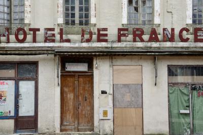 photograph “V.2015 — Victim of its own success” par David Farreny — www.farreny.net — France, bâtiment, building, façade, facade, hôtel, hotel, hôtel de France, Jacques Attali, Michel Houellebecq, abandonné, abandoned, abandon, enseigne, sign, lettres, letters, métal, metal, condamné, boarded up, fenêtres, windows, porte, door, restaurant, vitrines, rideaux, curtains, bois, wood, verre, glass, Occitanie, Gascogne, Gers, Gimont