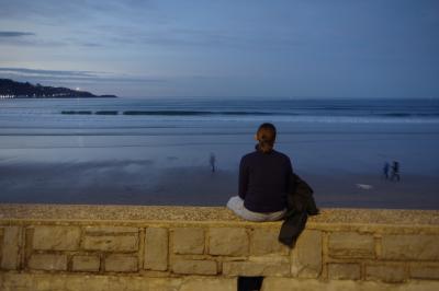 photograph “XII.2020” par David Farreny — www.farreny.net — France, Aquitaine, Pyrénées-Atlantiques, Hendaye, Pays basque, Basque country, océan, Atlantique, Atlantic, ocean, mer, sea, eau, water, paysage, landscape, marin, seascape, vagues, waves, ciel, sky, soir, evening, hiver, winter, bleu, blue, sable, sand, plage, beach, horizon, ligne, line, jeune, femme, young, woman, Lu*, assise, sitting, muret, low wall, pierres, stones, contemplation, piétons, pedestrians, flous, blurred, chien, dog, lumières, lights, rivage, seashore, carte postale, postcard