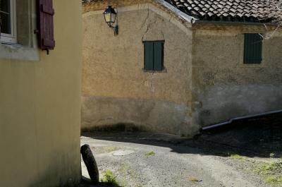 photograph “II.2019 — Faces of home, 145” par David Farreny — www.farreny.net — France, Occitanie, Gascogne, Gers, Montesquiou, maison, house, pierre, stone, fenêtres, windows, volets, shutters, bois, wood, ruelle, alley, rue, street, bitume, blacktop, asphalte, asphalt, lampe, lamp, toit, roof, tuiles, tiles, génoise, campagne, countryside, tuyau, pipe, branches, angle, coin, corner