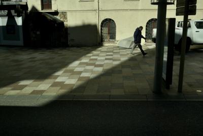 photograph “XI.2023 — Exit from the circus” par David Farreny — www.farreny.net — Andorre, Andorra, Escaldes-Engordany, ville, city, rue, street, trottoir, pavement, dallage, paving, pierre, stone, bâtiment, immeuble, building, passant, passer-by, piéton, pedestrian, homme, man, voiture, car, véhicule, vehicle, pick-up, métal, metal, poteaux, posts, poles, fenêtres, windows, grilles, railings, bitume, blacktop, asphalte, asphalt, ombres, shadows