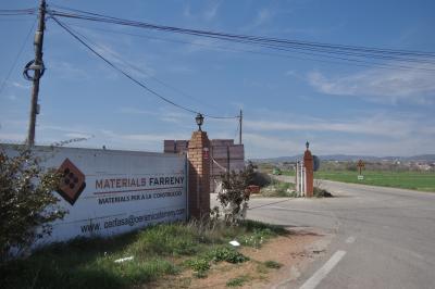 photograph “III.2023 — Miserable materialism” par David Farreny — www.farreny.net — Espagne, España, Spain, Catalogne, Cataluña, Catalunya, Lérida, Lleida, Balaguer, route, street, bitume, blacktop, asphalte, asphalt, poteau, post, pole, electricity, électrique, entreprise, company, câbles, cables, fils, wires, enseigne, sign, Materials Farreny, materials per a la construcció, clôture, fence, brique, brick, lampes, lamps, arbustes, shrubs, herbe, grass, champ, field, plaine, plain, tas, heaps, collines, hills, voiture, car, verte, green, panneau, routier, roadsign, ciel, sky, nuages, clouds
