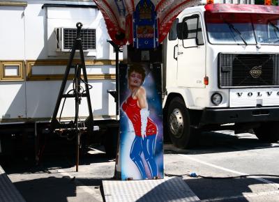 photograph “VI.2008 — Desire” par David Farreny — www.farreny.net — France, Occitanie, Aveyron, Rouergue, Rodez, fête foraine, funfair, camion, truck, lorry, Volvo, attraction, punching-ball, peinture, painting, femme, woman, laid, ugly, laideur, ugliness, caravane, caravan, trailer, van, métal, metal, parking, car park, bitume, blacktop, asphalte, asphalt, ombres, shadows, gyrophares, rotating lights