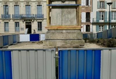 photograph “III.2024 — Death of the war memorial” par David Farreny — www.farreny.net — France, Occitanie, Tarn-et-Garonne, Caussade, monument aux morts, war memorial, barrières, barriers, métal, metal, clôture, fence, pierre, stone, chantier, work site, bois, wood, sol, soil, terre, earth, bâtiments, buildings, brique, brick, fenêtres, windows, volets, shutters, lampadaire, lamp, balcons, balconies, planches, planks, passante, passer-by, piétonne, pedestrian, femme, woman, vieille, old, béret, beret, socle, base, noms, names, soldats, morts, dead, soldiers, oubli, oblivion