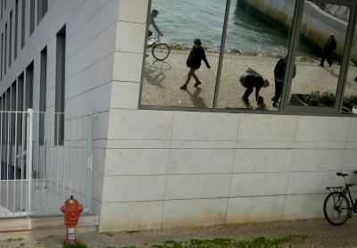 photograph “II.2020” par David Farreny — www.farreny.net — Portugal, Lisbonne, Lisboa, Lisbon, ville, city, capitale, bâtiment, building, béton, concrete, fenêtres, windows, baies, verre, vitre, glass, miroir, mirror, angle, coin, corner, clôture, fence, grille, railings, borne à incendie, hydrant, rouge, red, métal, metal, vélo, bicyclette, bicycle, bike, passants, passers-by, piétons, pedestrians, promeneurs, walkers, strollers, eau, water, fleuve, river, Tage, Tagus, quai, quay, rive, bank, rochers, rocks, pavés, trottoir, pavement, cobblestone, hiver, winter, herbe, grass