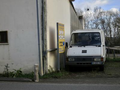 photograph “XII.2024 — Basic leisure” par David Farreny — www.farreny.net — France, Occitanie, Gascogne, Gascony, Lomagne, Tarn-et-Garonne, Beaumont-de-Lomagne, route, road, bitume, blacktop, asphalte, asphalt, bâtiment, building, béton, concrete, coin, angle, corner, fourgonnette, van, véhicule, vehicle, métal, metal, panneau, sign, base de loisirs, leisure center, entrée, entrance, flèche, arrow, droite, right, piscine, swimming pool, Le Lomagnol, logos, gourrière, drainpipe, poteau, post, pole, plantes, plants, arbres, trees, hiver, winter, tôle ondulée, corrugated iron, clôture, fence, verre, vitre, glass, branches, branchages, nuages, clouds, dépression, depression