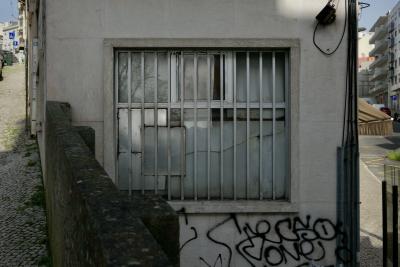 photograph “II.2020 — Cornershop” par David Farreny — www.farreny.net — Portugal, Lisbonne, Lisboa, Lisbon, ville, city, capitale, capital, immeubles, buildings, bâtiments, béton, concrete, coin, corner, angle, fenêtre, window, métal, metal, grille, railings, verre, vitre, glass, muret, low wall, trottoir, pavement, cobbling, panneaux, routiers, signs, roadsigns, voitures, cars, laideur, ugliness, laid, ugly, tags, graffitis, balcons, balconies, gouttière, drainpipe, câbles, cables, fils, wires, profusion, magasin, shop, store, commerce