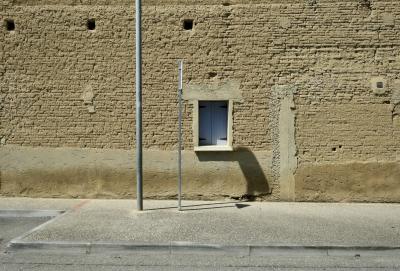 photograph “IV.2025 — Whatever” par David Farreny — www.farreny.net — France, Occitanie, Gascogne, Gascony, Lomagne, Tarn-et-Garonne, Beaumont-de-Lomagne, rue, street, trottoir, pavement, mur, wall, brique crue, raw brick, fenêtre, window, volets, shutters, post, pole, poteau, panneau, sign, ombres, shadows, métal, metal, bitume, blacktop, asphalte, asphalt, trous, holes