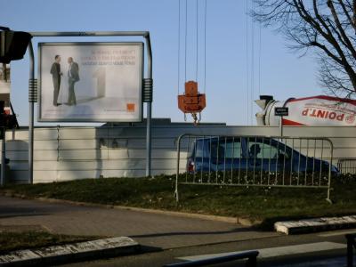 photograph “II.2012 — Satisfied when you are” par David Farreny — www.farreny.net — France, chantier, construction, building, site, palissade, boarding, arbre, tree, voiture, car, barrières, barriers, camion, truck, lorry, malaxeur, toupie, cement mixer, béton, concrete, Point P, plaque, rue, street, sign, avenue, Victor Hugo, poète, poet, soir, evening, ville, city, profusion, îlot directionnel, traffic island, passage, piétons, pedestrian, zebra, crosswalk, métal, metal, câbles, cables, fils, wires, publicité, advertising, pub, ad, panneau, affichage, billboard, hoarding, Orange, femme, woman, conductrice, driver, tôle ondulée, corrugated iron, poulie, pulley, block, wire, hiver, winter, Occitanie, Aveyron, Rouergue, Rodez