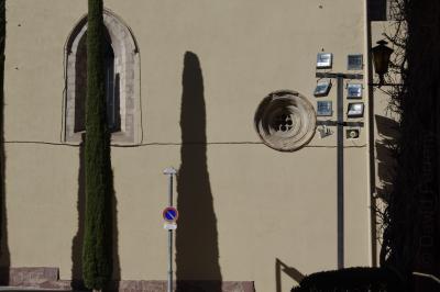 photograph “III.2023” par David Farreny — www.farreny.net — Espagne, España, Espanya, Spain, Catalogne, Cataluña, Catalunya, Lleida, Lérida, Alt Urgell, La Seu d'Urgell, ville, city, mur, paroi, wall, église, church, cyprès, cypress, ombre, shadow, gothique, gothic, fenêtres, windows, projecteurs, floodlights, poteau, post, pole, métal, metal, lampadaire, lamp, panneau, routier, sign, roadsign, câbles, cables, fils, wires, branches, hauts-parleurs, loudspeakers, gouttière, drainpipe, pierre, stone
