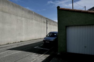 photograph “III.2022” par David Farreny — www.farreny.net — France, Occitanie, Ariège, Foix, rue, street, prison, maison d'arrêt, jail, mur, wall, béton, concrete, bitume, blacktop, asphalte, asphalt, voiture, car, garage, portail, gate, métal, metal, poteaux, posts, poles, câbles, cables, fils, wires, ombre, shadow, ciel, sky, tuiles, tiles