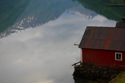 photograph “VI.2019 — Run away to the North, 157” par David Farreny — www.farreny.net — Norvège, Norway, Norge, Vestlandet, Sogn og Fjordane, Sogndal, Fjærland, fjord, eau, water, reflet, reflection, montagne, mountain, neige, snow, bâtiment, building, bois, wood, rouge, red, planches, planks, tôle ondulée, corrugated iron, toit, roof, toiture, roofing, rouille, rust, rusty, hangar, bateaux, boat, shed, pierre, stone, quai, rive, bank, métal, metal, été, summer, nuages, clouds, fenêtre, window, soir, evening