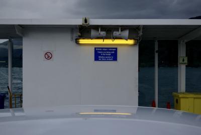 photograph “VI.2019 — Run away to the North, 278” par David Farreny — www.farreny.net — Norvège, Norway, Vestland, Sogn og Fjordane, Sogn, Høyanger, Lavik, Sognefjord, fjord, ferry, bateau, boat, métal, metal, voiture, car, toit, roof, néon, neon, lumière, lightning, haut-parleurs, loudspeakers, jaune, yellow, light, panneaux, signs, interdiction de fumer, no smoking, Les tickets sont vendus dans le salon, Billetter selges i salongen, Tickets are being sold in the lounge, Billetten werden in der Salon verkauft, reflet, reflection, nuages, clouds, gris, grey, gray, eau, water, montagne, mountain