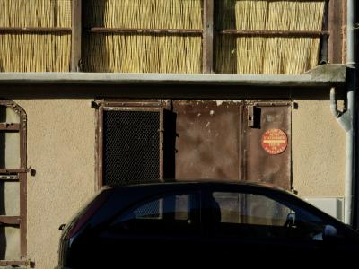 photograph “VI.2012” par David Farreny — www.farreny.net — France, rue, street, maison, house, façade, facade, voiture, car, métal, metal, panneau, roadsign, rideau, paille, curtain, straw, jaune, yellow, ciment, cement, gouttière, drainpipe, balcon, balcony, marron, brun, brown, grille, grate, portes, doors, été, summer, ville, city, Occitanie, Rouergue, Aveyron, Marcillac-Vallon