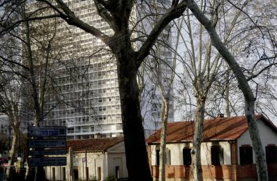 photograph “III.2024 — Faces of home, 215” par David Farreny — www.farreny.net — France, Occitanie, Haute-Garonne, Toulouse, Matabiau, immeuble, bâtiment, building, béton, concrete, fenêtres, windows, reflet, reflection, maisons, houses, arbres, trees, platanes, plane trees, branches, branchages, panneaux, signs, routiers, roadsigns, toits, roofs